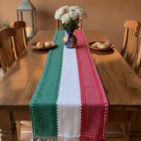 Camino de mesa tricolor tejido en algodón sobre mesa de madera con flores blancas y pan dulce, decoración mexicana tradicional.
