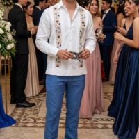 Esta camisa para hombre, estilo Guayabera o camisa de boda, es una prenda de vestir refinada y tradicional, ideal para ocasiones formales como bodas, graduaciones o celebraciones especiales. Confeccionada en un material que parece lino de alta calidad, ofrece una textura transpirable y elegante que proporciona comodidad sin perder el toque sofisticado. Detalles de Diseño: Bordado de Gala: El detalle más distintivo son las dos franjas verticales de bordado intrincado y detallado que recorren el frente de la camisa, a cada lado de la botonera. El diseño floral está realizado en hilos de color, creando un contraste rico y festivo. Aforzados Laterales: Para mantener la estética clásica de la guayabera, la camisa incluye aforzados (pliegues finos cosidos a mano) paralelos a los bordados, que añaden una textura sutil y elegante. Corte y Confección: Presenta un cuello cubano o cuello plano tradicional, sin abotonar el botón superior, ideal para un estilo "formal relajado". Las mangas son largas con puños sencillos. El corte es regular, ni demasiado ajustado ni muy holgado, para una silueta clásica.