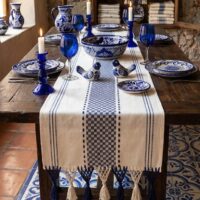 Camino de mesa artesanal en telar de pedal de Oaxaca, de color base crema/blanco hueso con patrones geométricos y franjas en azul cobalto estilo Talavera. La pieza presenta terminaciones de flecos artesanales anudados a mano