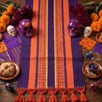 "Camino de mesa artesanal oaxaqueño de telar de pedal para Día de Muertos. Textil morado con franjas naranjas y amarillas, y flecos decorativos, usado como base para un altar tradicional